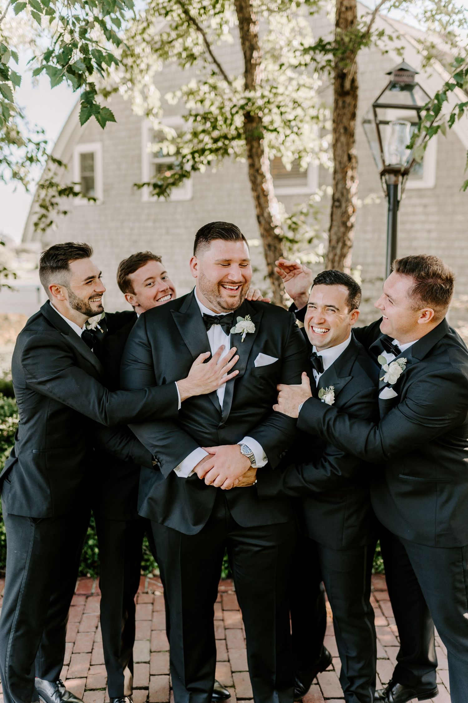 Groomsmen in black tuxedos share playful candid moments during outdoor wedding photos.