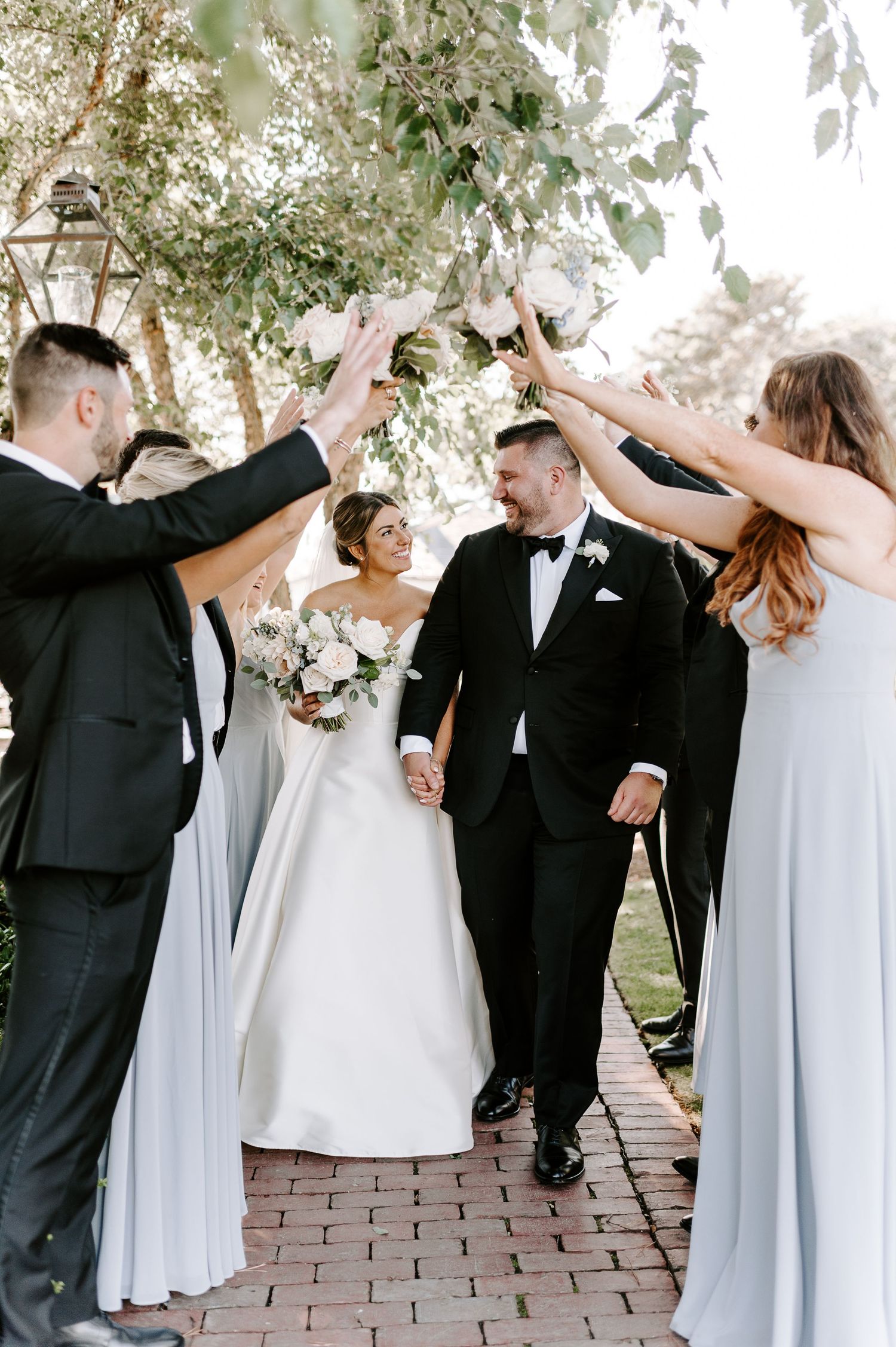 Wedding party celebrates with raised arms and joyful poses in outdoor garden setting.