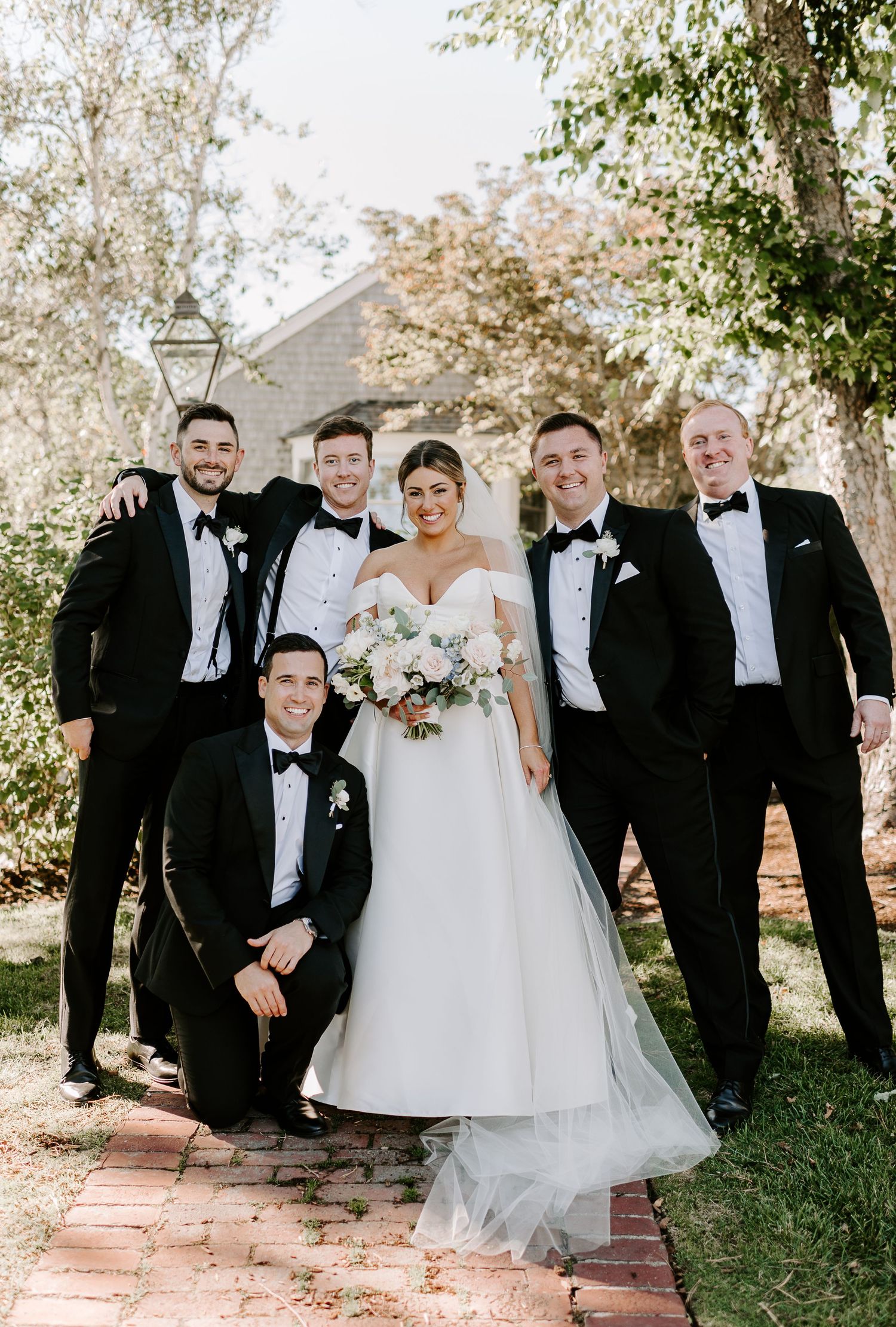 Wedding party in formal attire poses together on brick pathway surrounded by trees.