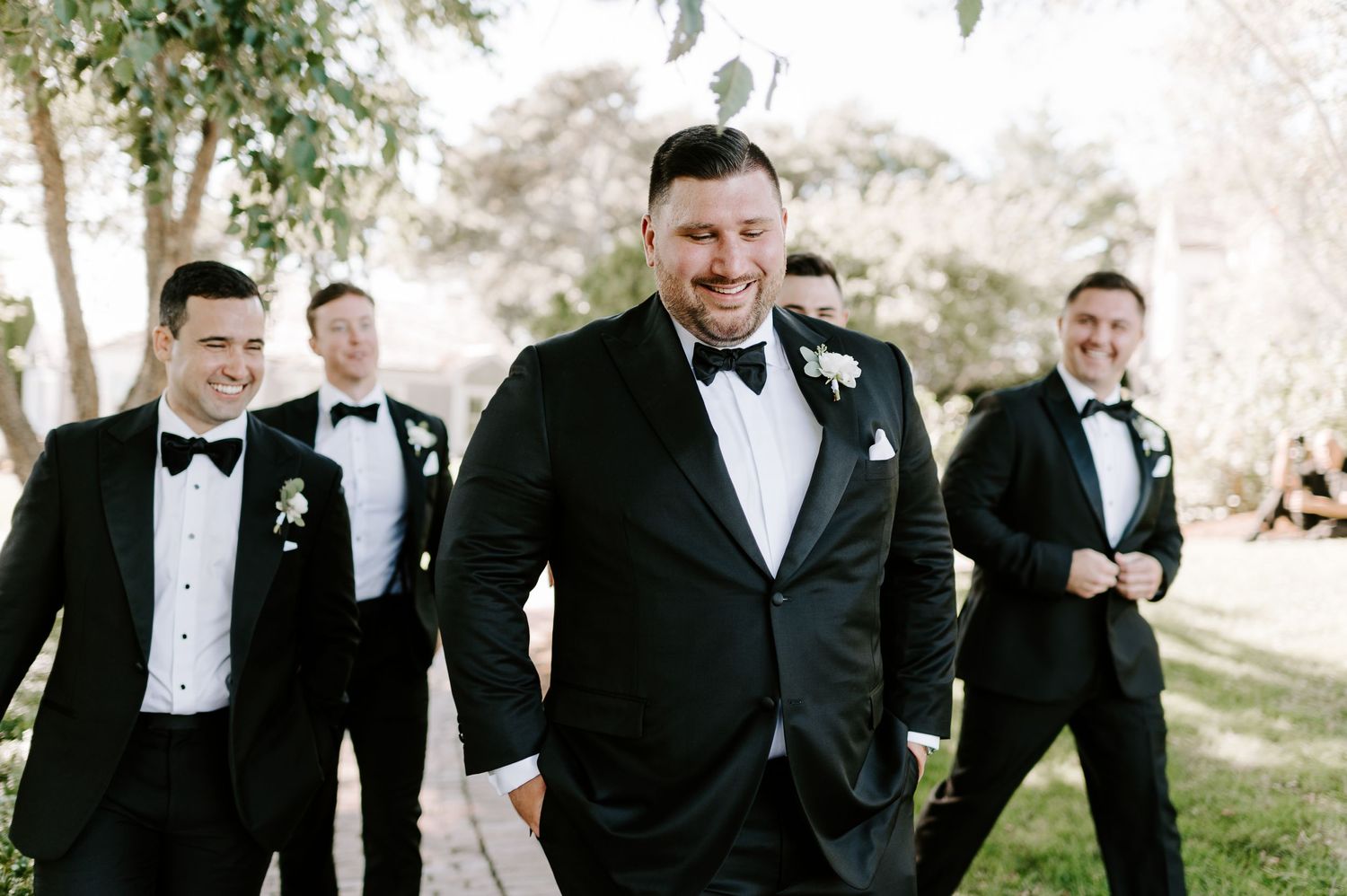 Groomsmen in black tuxedos share laughs while walking together outdoors.