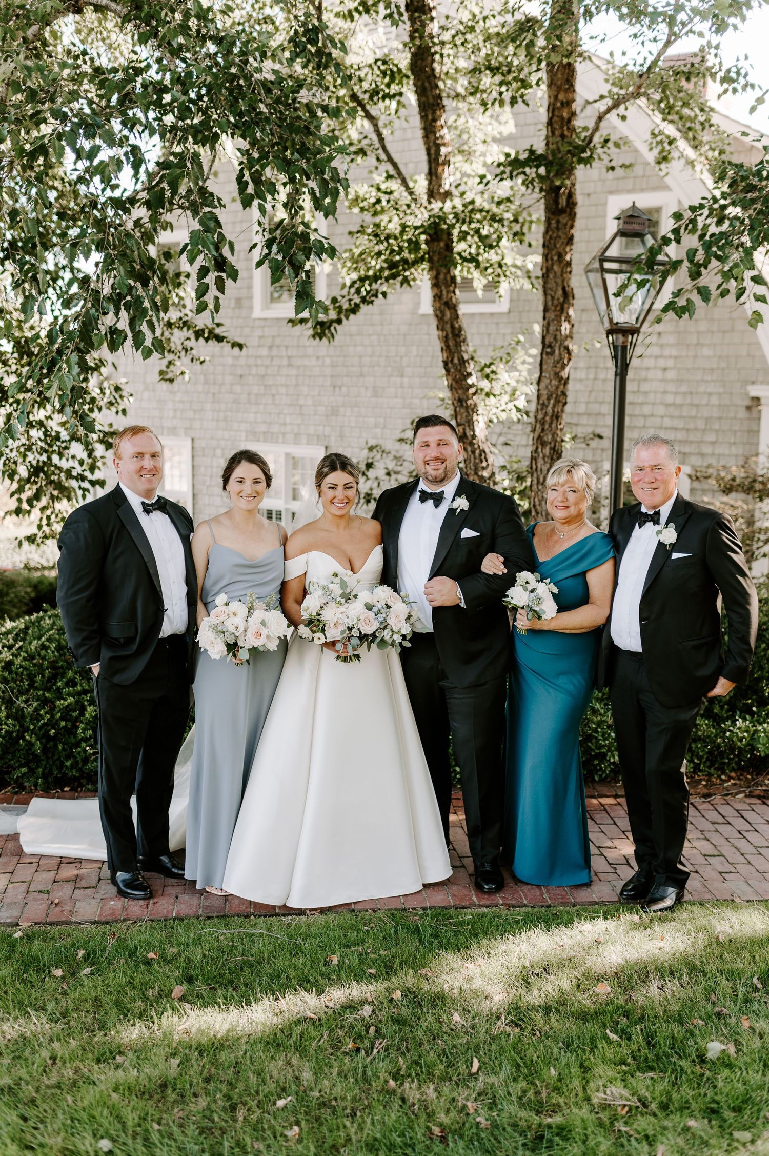 Wedding party in black tuxedos and formal dresses poses for photos on garden grounds.