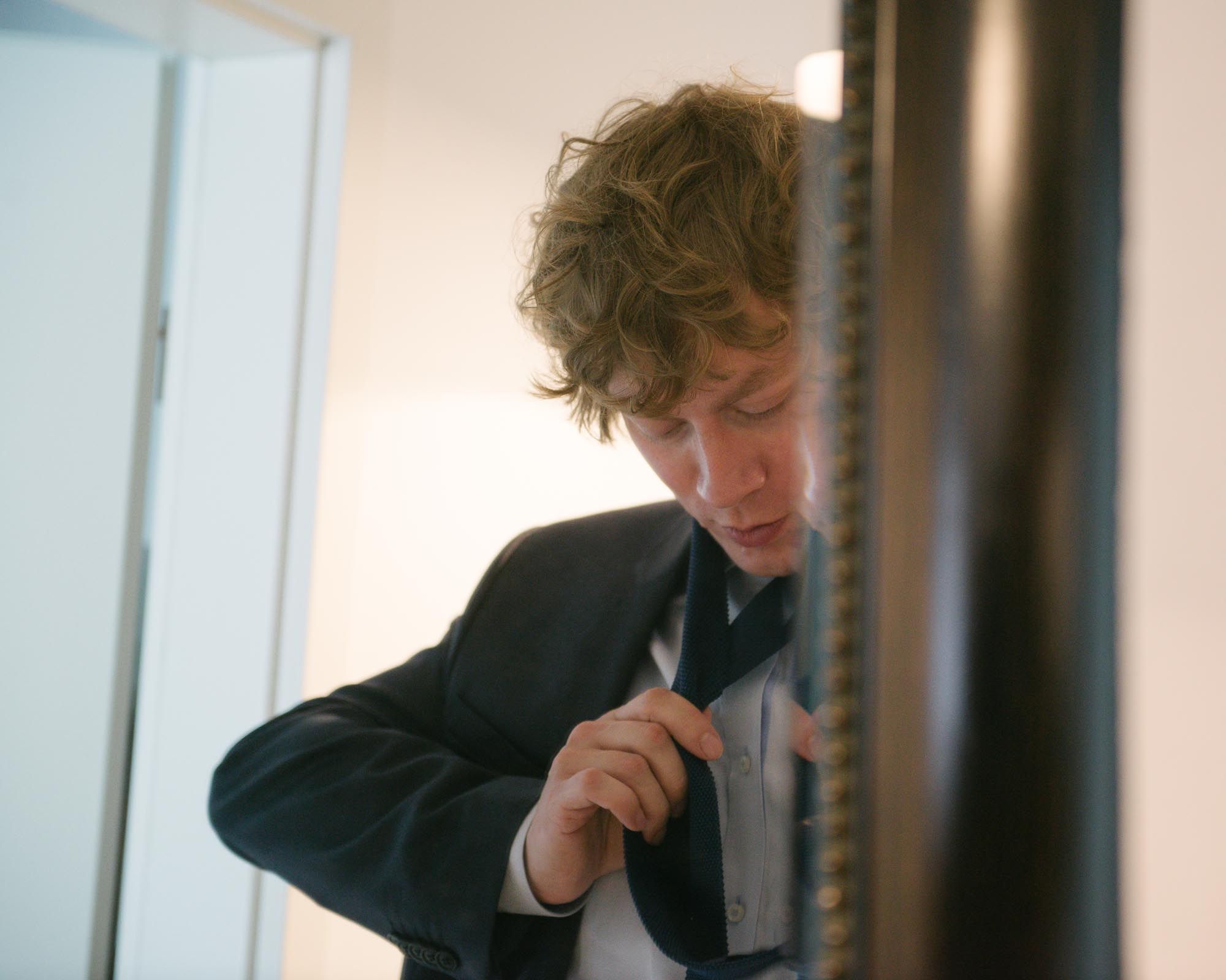 A well-dressed person in a dark suit adjusting their tie while looking down.