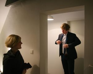 Two people in formal attire having a conversation in a minimalist white room.