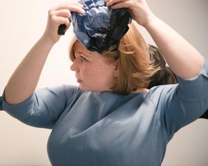 Someone in a blue dress adjusting a dark fascinator headpiece.