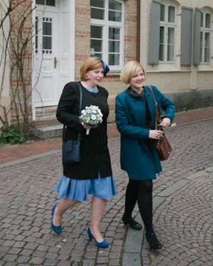 Two people in winter coats walking together on a cobblestone street, one carrying a white bouquet of flowers.