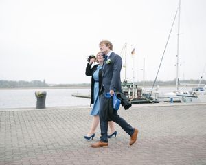 A couple walking along a harbor walkway in formal attire on an overcast day.