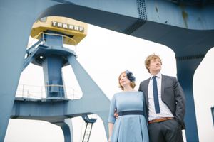 A couple poses near industrial maritime equipment on a boat deck with the woman wearing a blue vintage-style dress.