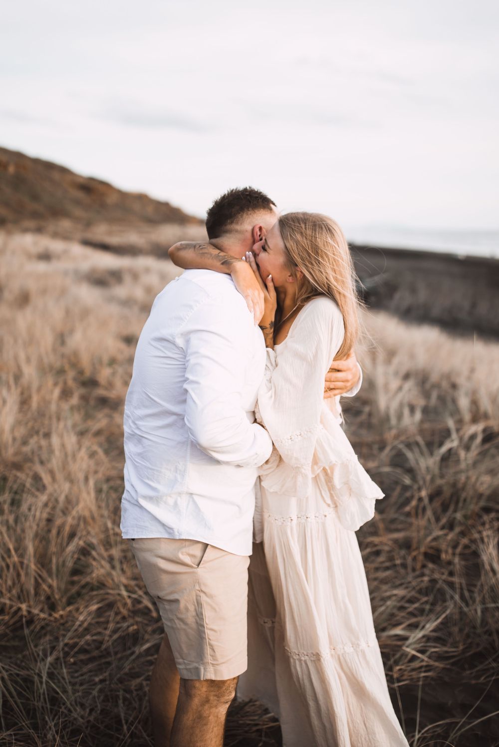 A romantic couple embraces in a field of tall grass during golden hour sunset.