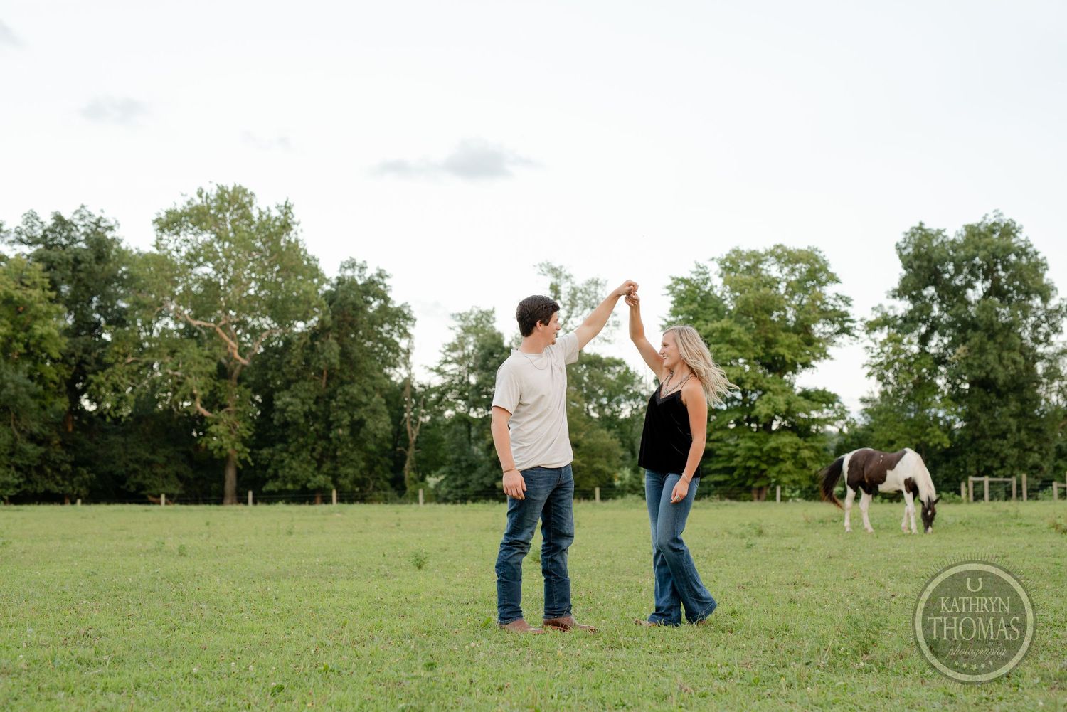 A playful couple dances in a summer field with horses in the distance.