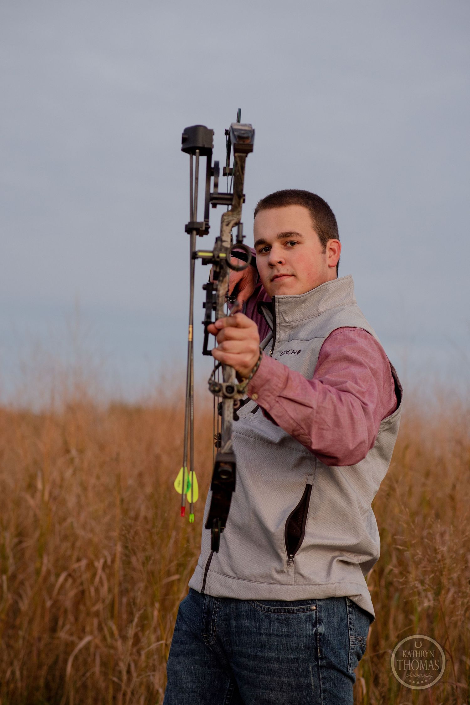 Archer aims modern compound bow in field with tall grass.
