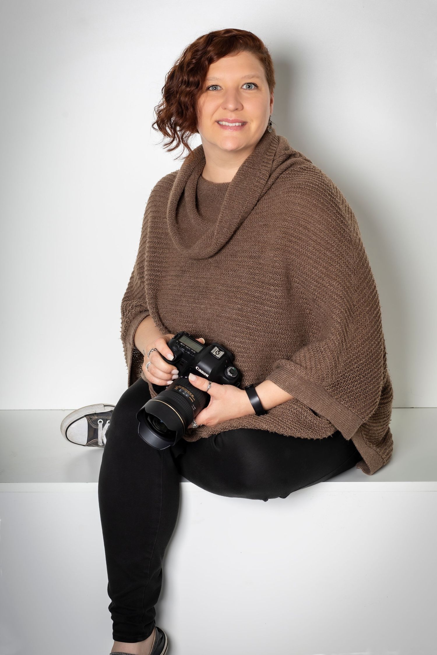 Professional photographer in brown poncho sweater holding camera in studio setting.
