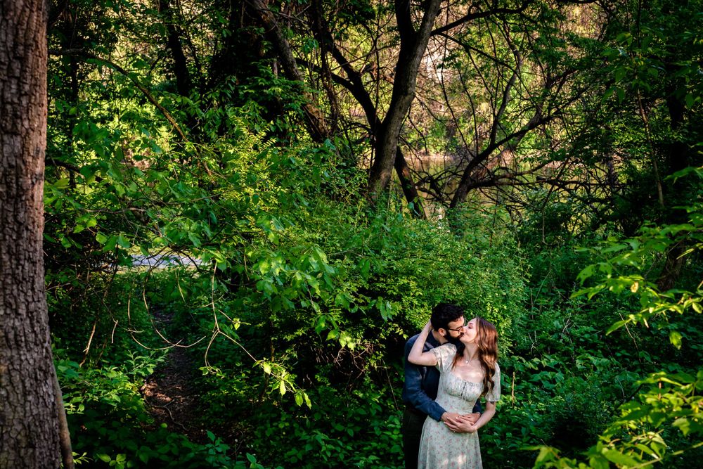 A couple embraces in a scenic wooded area with sunlight filtering through trees.