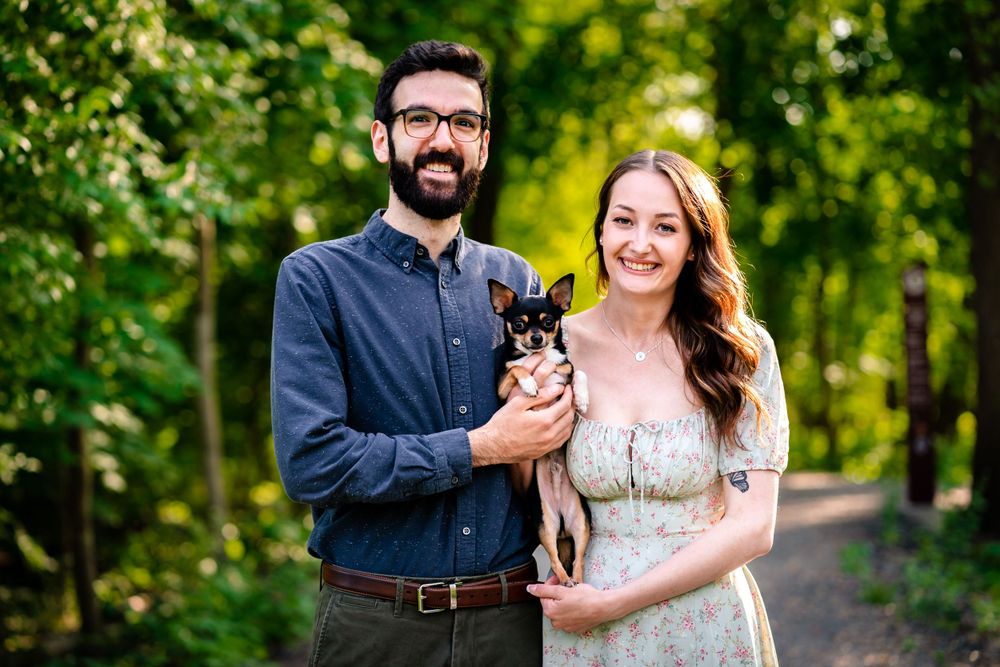 A couple poses with their small chihuahua dog on a tree-lined path.
