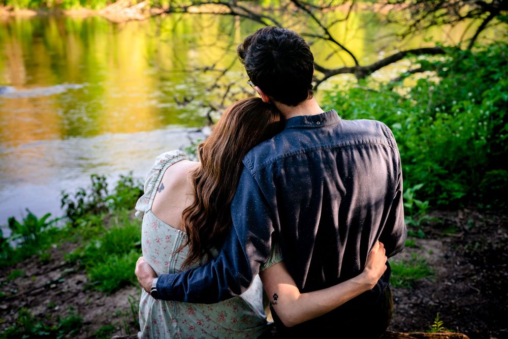 A couple stands by a reflective lake surrounded by green foliage.