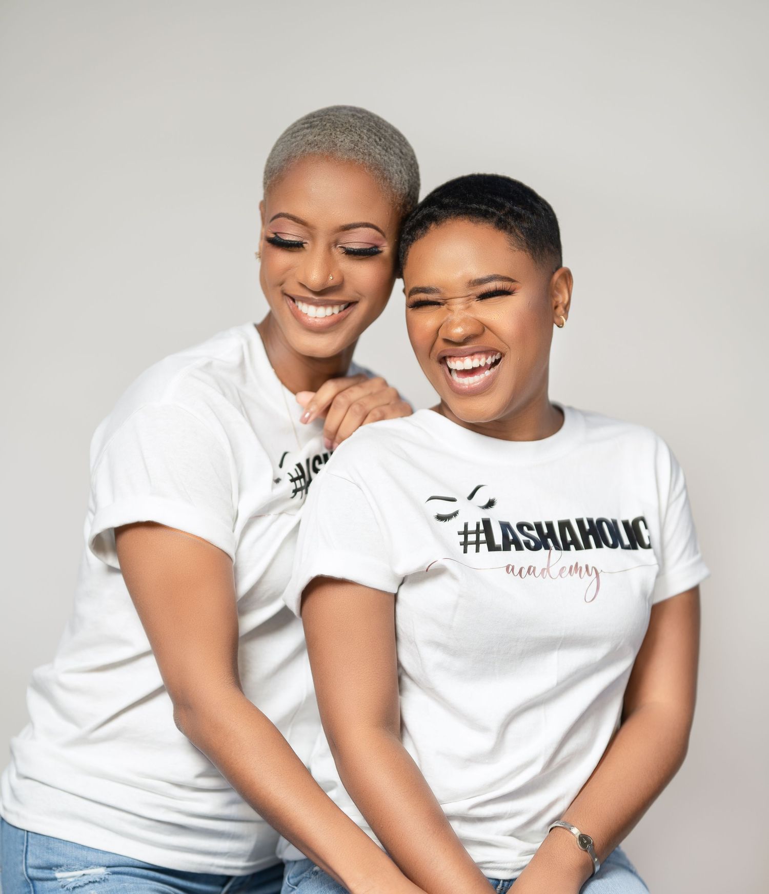 Two friends in matching white t-shirts sharing genuine laughter against studio backdrop.