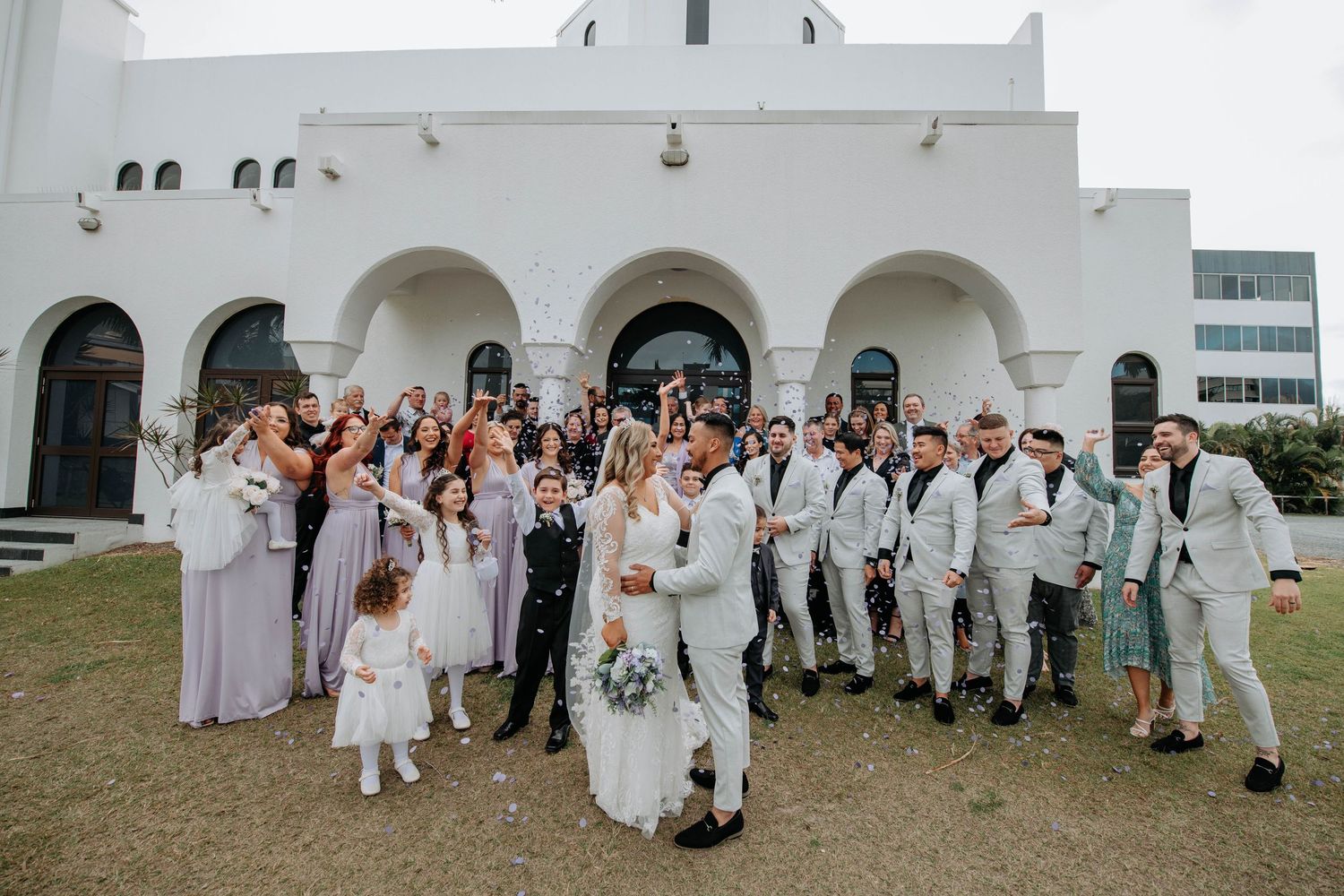Large wedding party poses together outside a white Spanish-style church building.