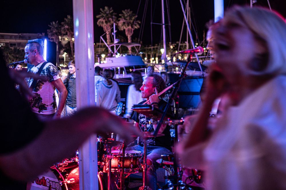 Drummer taking a break in a neon lit bar as party goers dance around him.