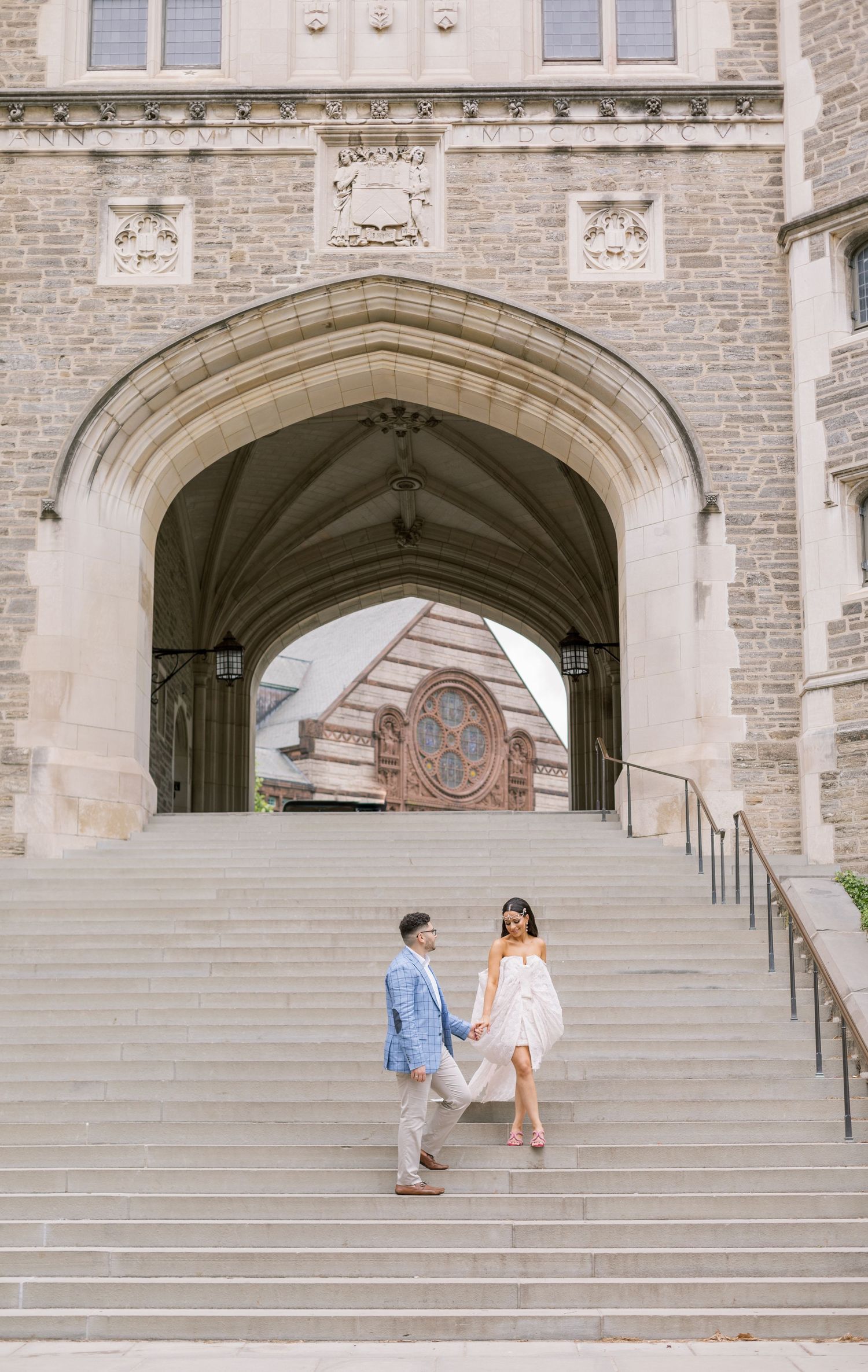 Couple in wedding attire poses on grand stone steps beneath Gothic archway.