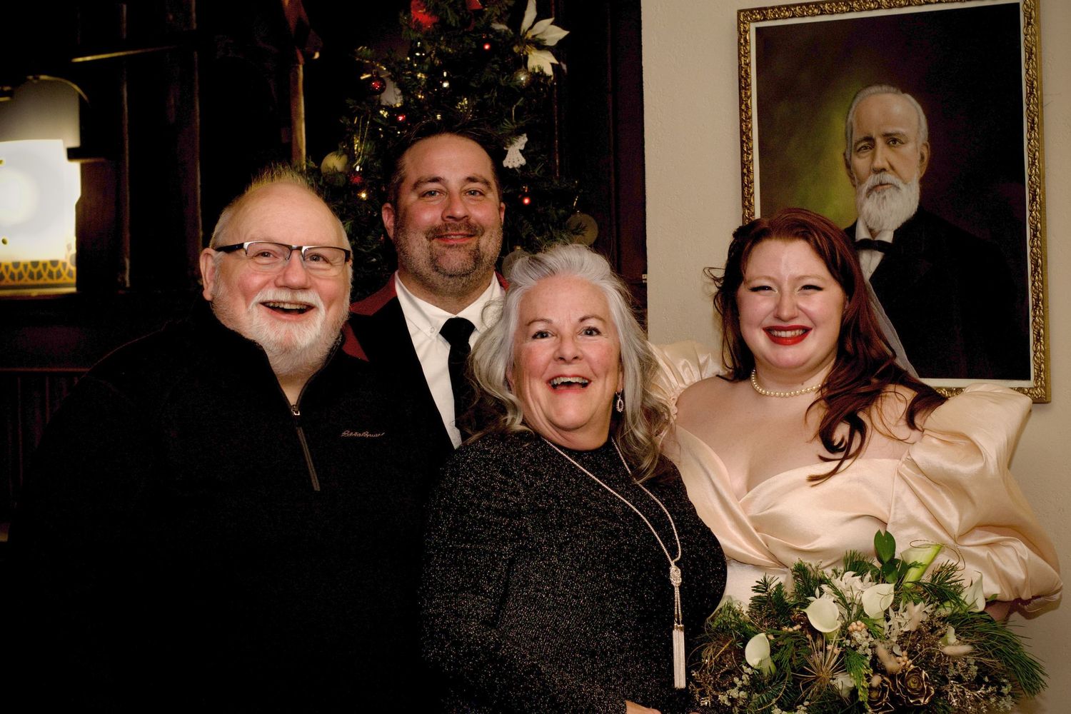 Four people pose together smiling at a wedding celebration indoors, with the bride holding a floral bouquet.