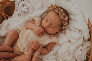 Sleeping newborn baby wearing a floral headband, dressed in cream lace outfit, resting in a wicker basket with white knit blanket.