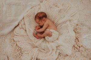 Newborn baby sleeping peacefully curled up on a cream crocheted blanket in a cozy, soft-lit nursery setting.