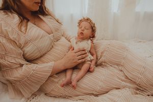 Mother in blush dress cradling sleeping newborn baby wearing floral crown in soft natural light.