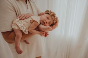 Adult cradling a sleeping newborn baby wearing a floral crown and lace bloomers against a white background.