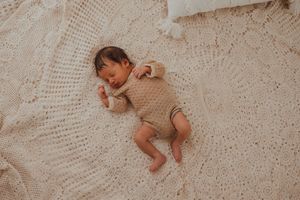 Baby sleeping peacefully on white lace blanket wearing beige knit romper.