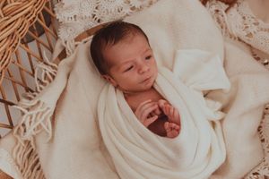 Sleeping newborn baby wrapped snugly in soft white swaddle blanket on textured cream macrame backdrop.