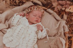 A sleeping infant in white knit clothing wears a floral crown against autumn-toned fabric.