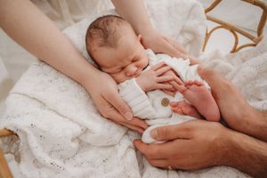 Newborn baby sleeping peacefully on white blanket while parents gently cradle and hold tiny feet.