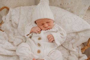 Newborn baby sleeping peacefully in a wicker basket, dressed in a white knit romper and matching beanie hat.