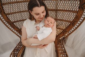 A mother in a white dress cradles her newborn baby while seated in a wicker peacock chair.