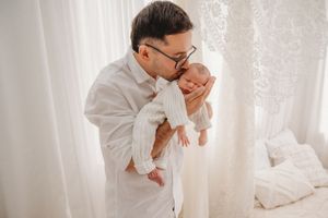 A father in a white shirt and glasses tenderly kisses his sleeping newborn baby in a bright, airy white room.