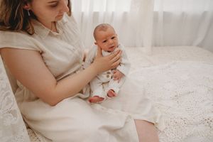 A mother in a cream dress tenderly holds her newborn baby in white clothing while seated on a lace-covered chair.