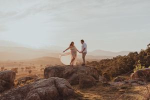Two figures hold hands while standing on a rocky cliff during golden hour.