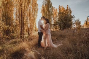 Couple embracing in golden autumn field surrounded by tall trees during romantic outdoor maternity photoshoot at sunset.