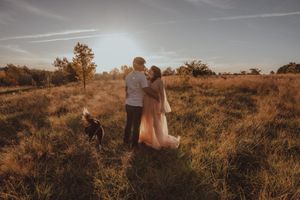 Couple standing in golden field at sunset with dog, wearing elegant pink dress, romantic outdoor photography.