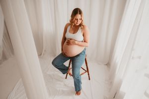 Pregnant woman in white crop top and jeans sitting on wooden stool, cradling baby bump in bright white studio.