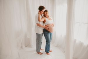 Couple sharing tender maternity moment, man embracing pregnant woman in white airy studio setting.