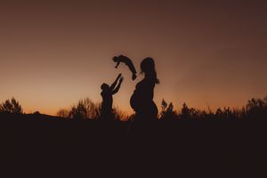 Silhouette of child and pregnant woman dancing at sunset with trees in background.