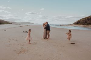 Couple sharing a romantic moment on a wide sandy beach while two young girls play nearby.