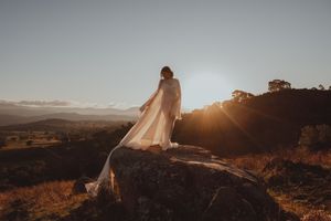Bride in flowing white gown standing on a rock at golden sunset with mountain landscape backdrop.