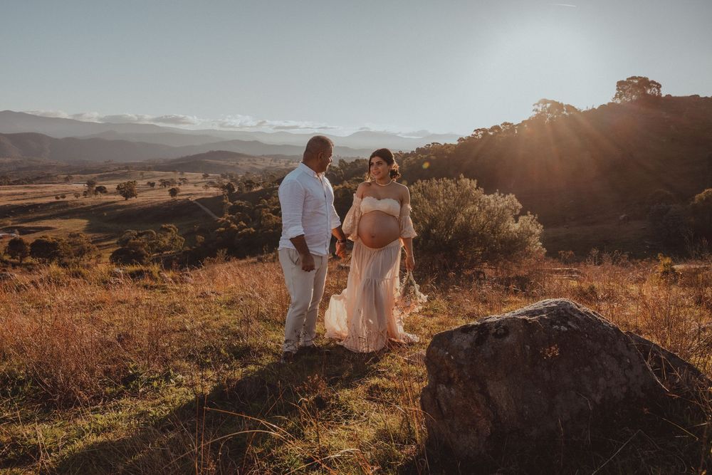 Couple sharing a tender maternity moment at golden hour on a scenic hilltop with mountains in the background.