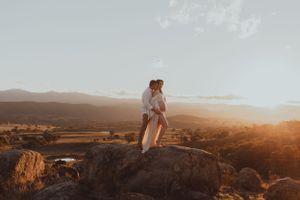 Couple embraces on rocky hilltop during golden hour in scenic mountain landscape.