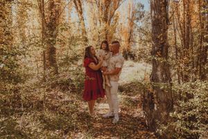 Family of three posing in autumn forest, parents holding toddler among golden trees with fallen leaves.