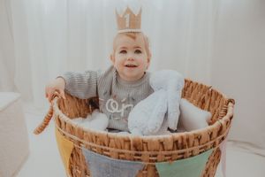 A smiling baby wearing a gold crown sits in a wicker basket with a stuffed bunny, celebrating their first birthday.