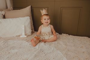 Baby in crown and ONE romper sits smiling on white crochet blanket during first birthday photo shoot.