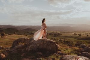 Pregnant woman in flowing pink gown stands on rock overlooking lush green valley at golden hour.
