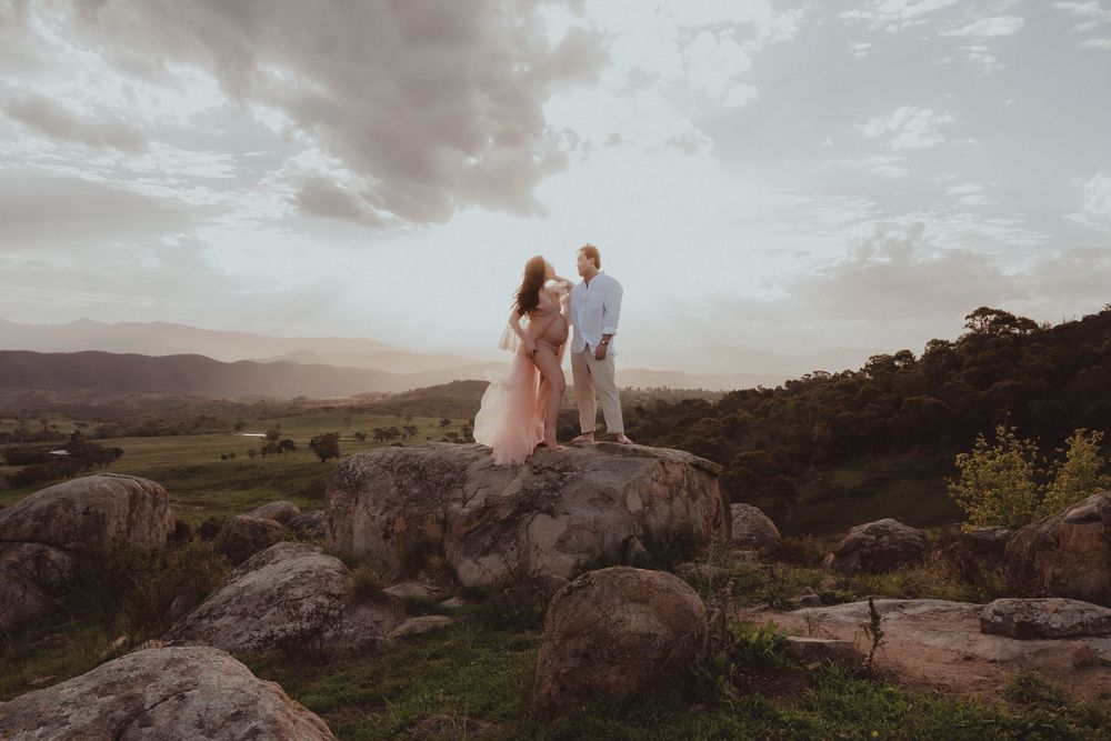 Couple standing on rocks at sunset with scenic mountain valley and dramatic cloudy sky in background.
